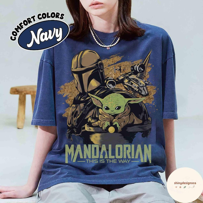 Puede incluir: Camiseta azul marino con un dise&ntilde;o de The Mandalorian y Grogu, una nave espacial y el texto "MANDALORIAN - THIS IS THE WAY." La camiseta tiene una etiqueta Comfort Colors en un &oacute;valo azul marino.