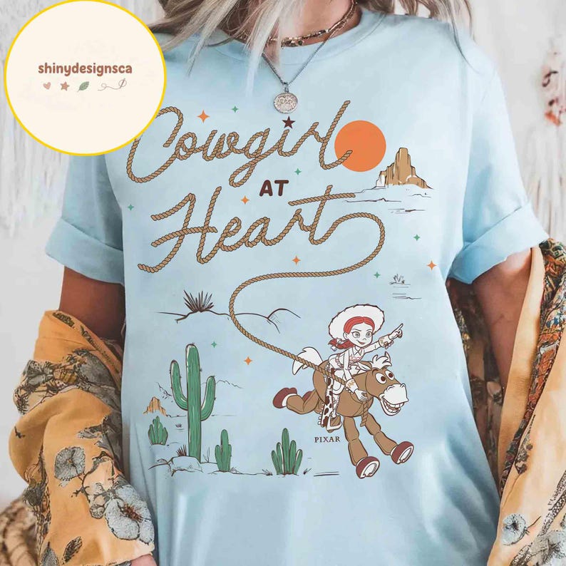 May include: Light blue t-shirt with the text "Cowgirl at Heart" in a rope-like font. The shirt features a cartoon illustration of a cowgirl riding a horse, with a desert landscape and a setting sun in the background. The word "Pixar" is also visible.