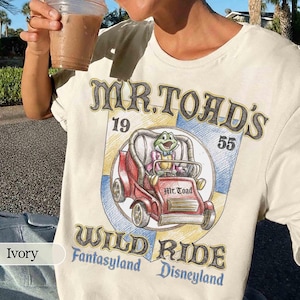 Pode incluir: Camiseta de cor marfim com um gráfico de Mr. Toad num carro vermelho, com o texto "Mr. Toad's Wild Ride", "Disneyland" e "Fantasyland". O design inclui os números "19" e "55".