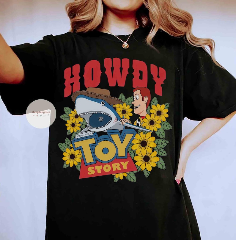 Puede incluir: Camiseta negra con un dise&ntilde;o gr&aacute;fico. El dise&ntilde;o presenta la palabra "HOWDY" en rojo, un tibur&oacute;n con sombrero de vaquero, Woody de Toy Story, girasoles y las palabras "TOY STORY" en amarillo y azul.