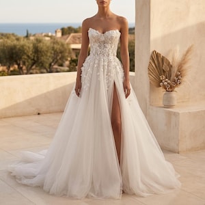 Könnte beinhalten: Ein trägerloses weißes Hochzeitskleid mit floralen Verzierungen und einem hohen Schlitz. Das Oberteil hat aufwendige Spitzendetails, und der Rock geht in eine lange Schleppe über. Das Kleid wird im Freien mit einem malerischen Hintergrund getragen.