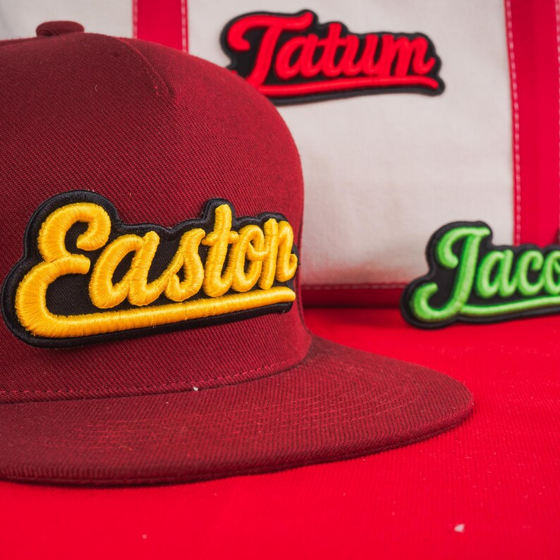 May include: A maroon baseball cap featuring the name Easton embroidered in yellow cursive script with a black outline. In the background, a white canvas bag displays the names Tatum in red and Jacob in green, both in cursive script.