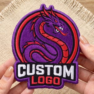 May include: A circular embroidered patch featuring a purple dragon with red accents coiled inside a red ring. Below the dragon, a black rectangular base displays the white text CUSTOM and the red text LOGO, all bordered by a purple stitched edge.