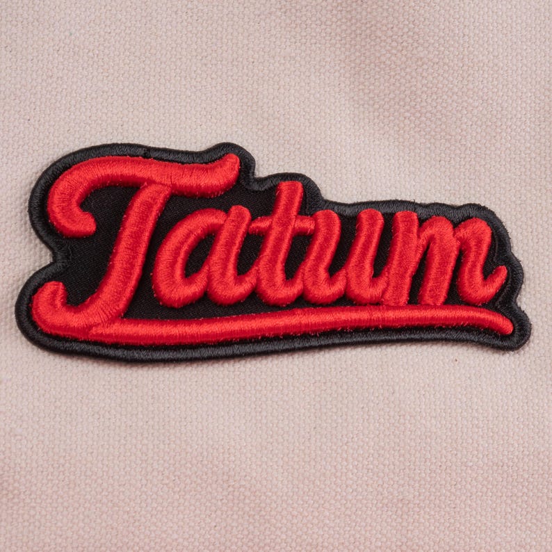 May include: An embroidered patch featuring the word Tatum in a bold, red, cursive script font. The lettering is outlined in black thread and set against a black fabric background, creating a distinct contrast against the light beige textile base.