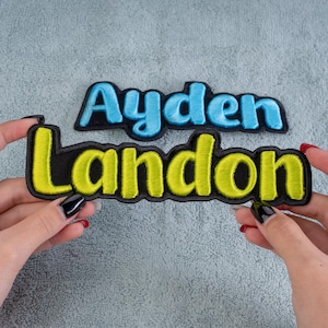 May include: Two embroidered patches with names. One patch says "Ayden" in light blue letters with a black border. The other patch says "Landon" in yellow letters with a black border. Both patches are on a gray surface.