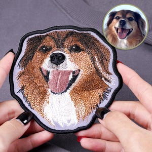 May include: Embroidered patch featuring a smiling dog with brown and white fur, a pink tongue, and a black outline. A smaller circular image of the dog is in the upper right corner. The patch is held by hands with manicured nails.