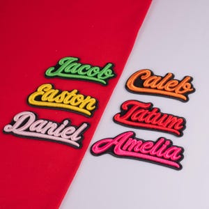 May include: Six embroidered name patches with black borders and cursive lettering. The names Jacob, Easton, and Daniel are in lime green, yellow, and light pink on a red fabric background. Caleb, Tatum, and Amelia are in orange, red, and hot pink.