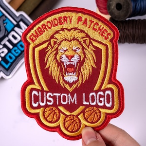 May include: A shield-shaped embroidered patch featuring a gold lion head on a dark red background. The text EMBROIDERY PATCHES is arched at the top, and CUSTOM LOGO is written in white below the lion. Three basketballs are depicted at the bottom.