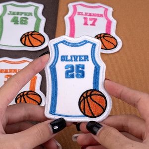 May include: White basketball jersey patches with blue, green, orange, and pink trim. Each patch features a name and number, such as "Oliver 25" and "Jesper 46", along with a basketball graphic. The patches are embroidered.