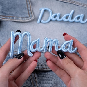May include: Light blue embroidered patches spelling "Mama" and "Dada" on a denim jacket. The patches are in a cursive font and are made of a soft, textured material. The background is a light blue denim.