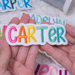 May include: A white embroidered name patch featuring the name CARTER in colorful thread. The letters are stitched in pink, orange, yellow, teal, and blue, with a scalloped white border surrounding the text on a white fabric background.