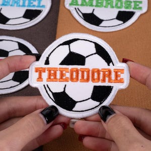 May include: Embroidered soccer ball patches with names. The patch in focus has the name "THEODORE" in orange letters. Other patches with names are visible. The patches have a white border and are on a brown background.