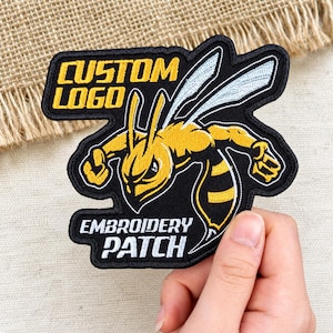May include: A black embroidered patch featuring a yellow hornet mascot with white wings. The text CUSTOM LOGO is embroidered in yellow at the top, and the text EMBROIDERY PATCH is embroidered in white at the bottom of the patch.