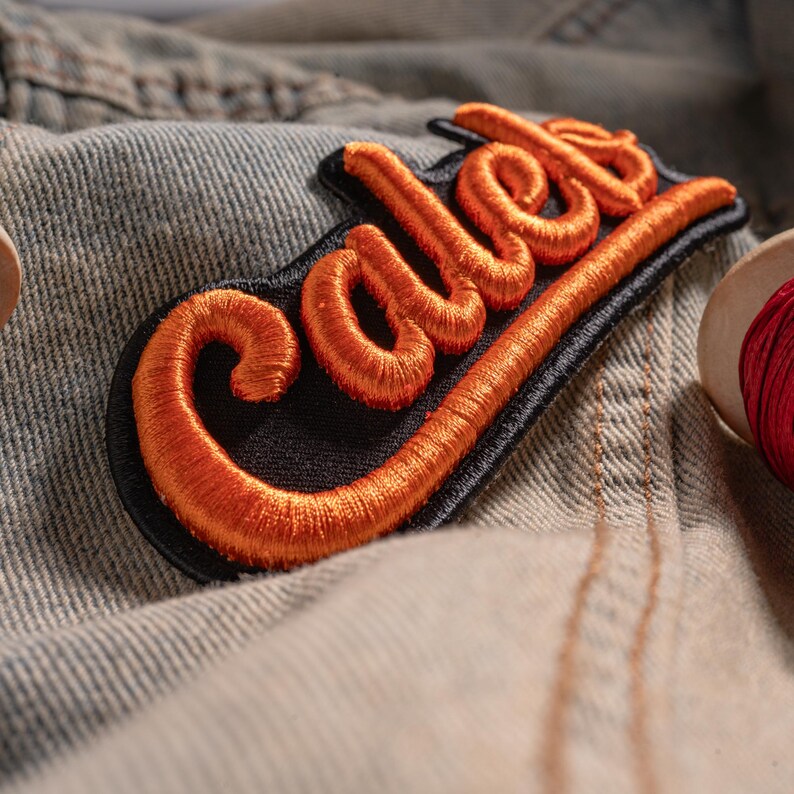 May include: An embroidered patch featuring the name Caleb in raised, bright orange thread with a thick, rounded script font. The lettering is outlined and set against a solid black fabric backing, creating a bold contrast for the name design.