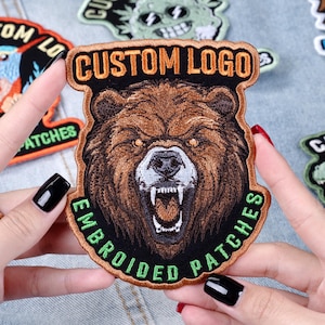 May include: Embroidered patch featuring a detailed brown bear head with an open mouth, baring its teeth. The patch has the text "CUSTOM LOGO" at the top and "EMBROIDERED PATCHES" at the bottom, with a black and brown border.