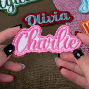May include: A pink embroidered patch with the name "Charlie" in a cursive font, held between two hands. Other patches with names are visible in the background, including "Olivia" and "ty". The patches have a white background and colorful borders.