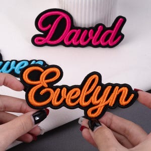 May include: Embroidered name patches in pink, orange, and blue script lettering on black fabric. The name "David" is in pink, and "Evelyn" is in orange. The patches are likely for clothing or accessories.