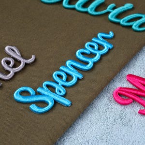 May include: Close-up of embroidered name patches in various colors. The name "Spencer" is prominently displayed in a vibrant blue, with other names in teal, pink, and lavender. The patches are on a brown and light blue textured surface.