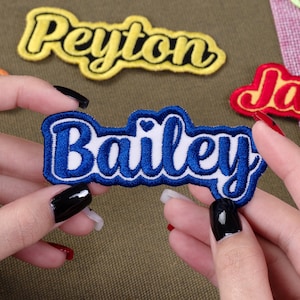 May include: A blue and white embroidered patch with the name "Bailey" in a cursive font, held between two hands. The patch has a blue border and a small heart detail. Other patches with names are visible in the background.