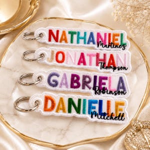 May include: Four white embroidered fabric keychains with silver metal rings. Each keychain features a first name in colorful block letters and a last name in black cursive script: Nathaniel Martinez, Jonathan Thompson, Gabriela Robinson, and Danielle Mitchell.