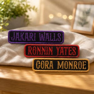 May include: Three rectangular embroidered name patches with black fabric backgrounds. The top patch features purple text reading JAKARI WALLS, the middle patch has red text reading RONNIN YATES, and the bottom patch has gold text reading GORA MONROE.