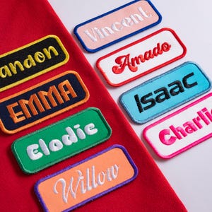 May include: A collection of rectangular embroidered name patches in various colors including red, black, navy, green, orange, light blue, pink, and white. The patches feature names such as Brandon, Emma, Elodie, Willow, Vincent, Amado, Isaac, and Charlie.