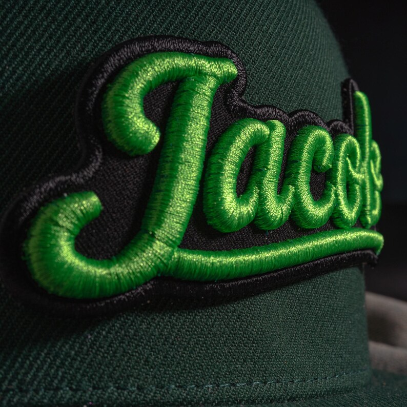 May include: A dark green baseball cap featuring a raised, embroidered patch with the word "Jacobs" in bright lime green script lettering outlined in black. The embroidery has a textured, three-dimensional appearance against the fabric.