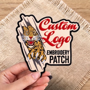 May include: An embroidered patch featuring a leopard head with bared teeth and claws emerging from a claw-mark design. The patch includes the red text Custom Logo and the black text EMBROIDERY PATCH on a cream-colored background.