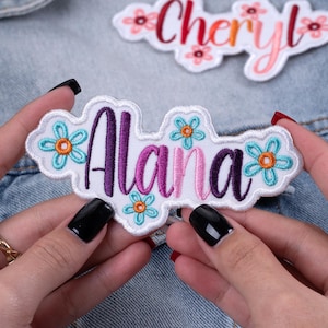 May include: Embroidered patch with the name "Alana" in purple and pink script, surrounded by blue and orange flower accents. The patch has a white border and is held against a denim background. Another patch with the name "Cheryl" is visible in the background.