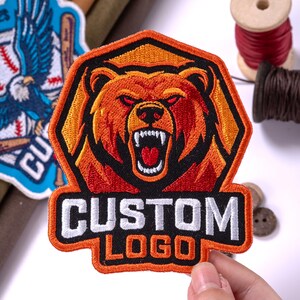May include: An embroidered patch featuring a stylized orange and red bear head with an open mouth and sharp teeth, set within a hexagonal border. Below the bear, the text CUSTOM LOGO is stitched in bold white and orange lettering.
