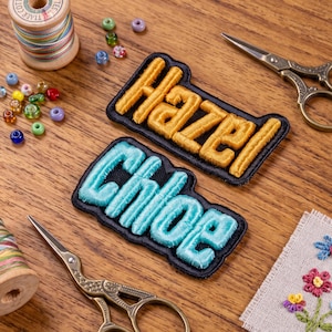 May include: Two rectangular embroidered patches with black borders, one featuring the name Hazel in raised gold thread and the other featuring the name Chloe in raised light blue thread, surrounded by colorful beads, thread spools, and scissors.