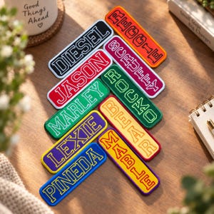 May include: A collection of rectangular embroidered name patches in various colors including black, red, green, purple, blue, orange, and yellow. Each patch features a bold, block-letter name stitched in a contrasting thread color.