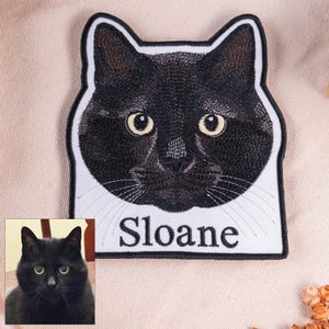 May include: An embroidered patch featuring the head of a black cat with yellow eyes and white whiskers. The patch has a white background, a black border, and the name Sloane embroidered in black text below the cat's chin. A small photo of the cat is included.