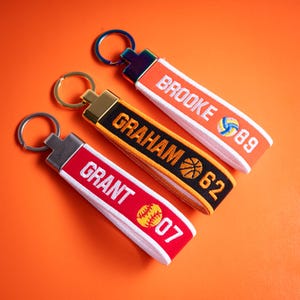 May include: Three fabric keychains with metal rings. The red one features the name GRANT, a yellow softball, and the number 07. The black one features the name GRAHAM, a basketball, and 62. The orange one features the name BROOKE, a volleyball, and 89.