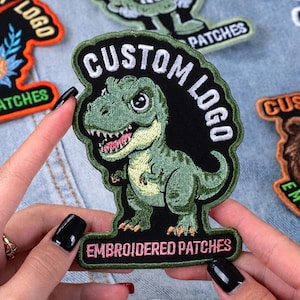 May include: Embroidered patch featuring a green cartoon Tyrannosaurus Rex with the text "CUSTOM LOGO" and "EMBROIDERED PATCHES" in white and pink. The patch is held against a denim background, showcasing its design and details.