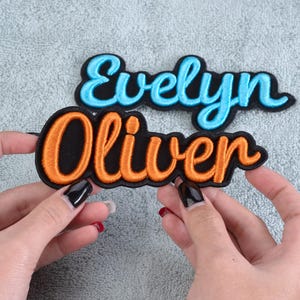 May include: Two embroidered name patches. One patch features the name "Evelyn" in light blue script with a black border. The other patch has the name "Oliver" in orange script with a black border.