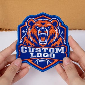 May include: An embroidered patch featuring a snarling orange bear head centered within a blue shield-shaped border. The text CUSTOM LOGO is stitched in white below the bear, with a small white football graphic at the bottom point.