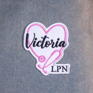 May include: An embroidered patch featuring a white heart outlined in pink thread. A pink stethoscope forms the heart shape. The name Victoria is stitched in black script across the center, with the letters LPN in black block text below.