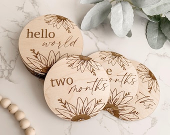 May include: A stack of round wooden baby milestone markers. Each marker features a sunflower design and text such as "hello world" and "two months". The markers are made of light-colored wood.
