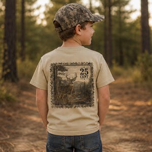 May include: A beige short-sleeved t-shirt featuring a square graphic on the back. The graphic depicts a deer in a forest landscape with the text 25 CENT. The shirt is paired with a camouflage baseball cap and blue denim jeans.