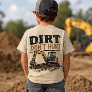 May include: A beige short-sleeve t-shirt featuring a graphic on the back with the text DIRT DON'T HURT in black and brown lettering above an illustration of a yellow excavator digging into a pile of dirt.