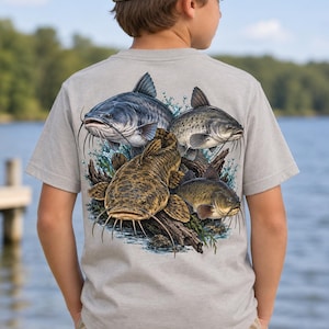 Puede incluir: Una camiseta gris claro de manga corta con un estampado gráfico detallado en la espalda. El diseño muestra cuatro siluros nadando entre troncos sumergidos y salpicaduras de agua. Se lleva con una gorra beige y pantalones de color caqui.