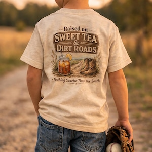 May include: A child wearing a tan t-shirt with the text "Raised on Sweet Tea & Dirt Roads" and a graphic of sweet tea and cowboy boots. The child is wearing blue jeans and a baseball cap, holding a baseball glove and ball.