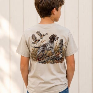 Puede incluir: Una camiseta beige de manga corta con un gráfico detallado en la espalda. La ilustración muestra un perro de caza moteado marrón y blanco de pie en hierba alta, rodeado de tres codornices volando y posadas en un entorno natural.
