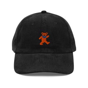 May include: Black corduroy baseball cap featuring an embroidered orange bear with a blue scarf. The cap has a curved brim and a top button. A classic, casual accessory.