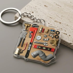 May include: A keychain featuring a collection of plumbing tools, including a red pipe wrench with the name "DAVID", pliers, and a level. The tools are arranged on a wooden background, with a silver chain and keyring.