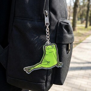 May include: A clear acrylic keychain featuring a green frog illustration with a bored expression, attached to a silver-toned metal chain and a split ring, currently clipped onto the zipper pull of a black fabric backpack.