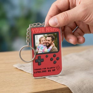 May include: Red acrylic keychain in the shape of a retro video game controller. The keychain features a photo of a father and child, with the text "YOUR NAME" and "THANKS FOR ALWAYS LEVELING US UP".