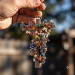 May include: A decorative acrylic ornament shaped like a utility pole with a lineman climbing it. The lineman wears a yellow hard hat with the name DAVID printed on it. The pole is wrapped in colorful Christmas lights in red, green, and yellow.