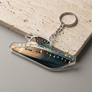 May include: A clear acrylic keychain featuring a boat design with colorful string lights. The keychain has a silver chain and ring. The boat is white and blue, with a detailed illustration of the boat's interior.